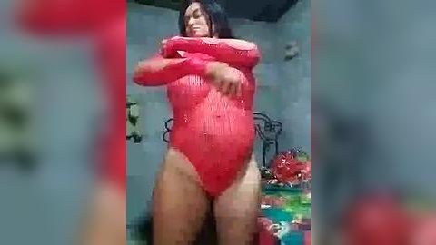 Media: Video of a woman in a red, wet, clingy bodysuit, standing in a dimly lit room with a bed and colorful objects in the background.