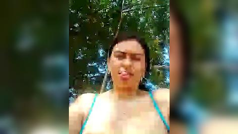 Media: A blurred video of a woman in a turquoise bikini standing in a lush, green forest setting. Her expression is playful, with her tongue sticking out. The background features trees and sunlight filtering through the foliage.