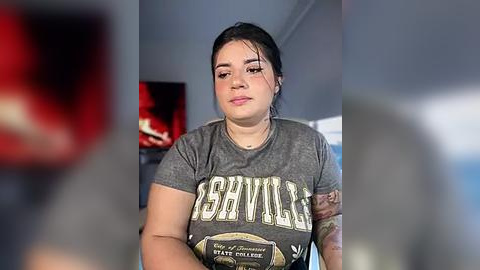 Media: Video of a young woman with light brown skin, dark hair, and tattoos on her arm, wearing a grey \"Ashville\" t-shirt, sitting indoors with blurred background.