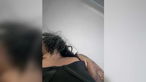 Media: Video of a person with short black hair and a colorful tattoo sleeve, wearing a black top, seen from behind. The background is a plain white wall.