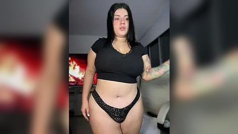 Media: Video of a curvy, plus-sized woman with long black hair, wearing a black crop top and lace panties, standing in a dimly lit room with a TV and blurred figures in the background.