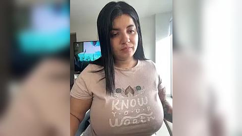 Media: Video of a Latina woman with long black hair, wearing a light pink t-shirt with \"Know Your Worth\" slogan. Background shows a TV with a blue screen.