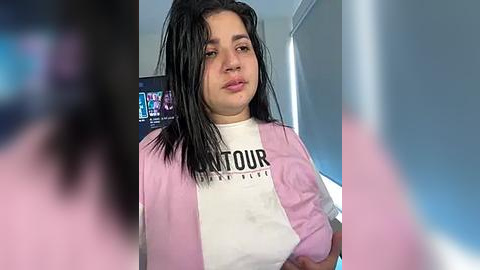 Media: Video of a young woman with long black hair, wearing a pink jacket over a white T-shirt with black text, standing indoors.