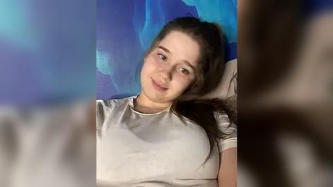 Media: Video of a young Caucasian woman with fair skin, brown hair tied back, and a casual grey t-shirt, lying on a bed with blue wall art in the background.