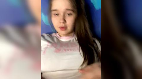 Media: Video of a young girl with long brown hair, wearing a white T-shirt with pink text, sitting in front of a blue wall. The image is blurred, with a focus on the girl's face.