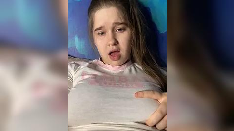 Media: Video of a young girl with light skin and long brown hair, wearing a light-colored shirt, lying on a blue pillow, looking shocked or in pain.