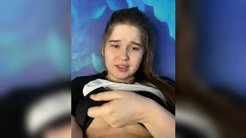 Media: Video of a young, fair-skinned woman with long brown hair, lifting her shirt to reveal her small breasts. Background features blue floral patterns.