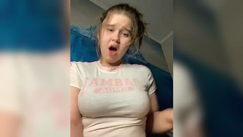 Media: A video of a young Caucasian woman with light skin, brown hair in a messy bun, wearing a white t-shirt with \"AMEN\" in pink, sitting on a blue couch, eyes closed, mouth open in apparent surprise or excitement.