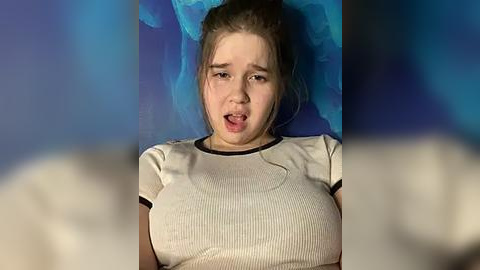 Media: A video of a young woman with light skin and brown hair, wearing a tight white shirt, lying on a blue background. Her mouth is open in apparent distress or surprise.