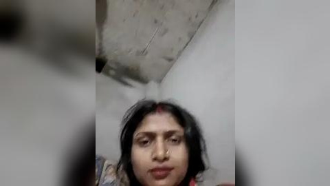 Media: Video of a South Asian woman with medium skin tone, wearing a red bindi, and black hair, standing in a dimly lit, small, white-walled room with a concrete ceiling.