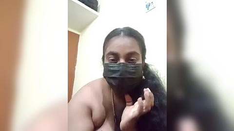 Media: Video of a topless Black woman with medium-dark skin tone, wearing a black mask and black bra, standing in a beige-walled room with a wooden door in the background.