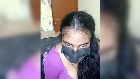 Media: Video of a woman with long black hair, wearing a purple top and black face mask, indoors near a door with peeling paint.