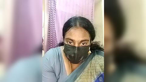 Media: Video of a middle-aged woman with medium brown skin and black hair, wearing a light blue saree, black mask, and a silver necklace, standing in front of a purple lace curtain.