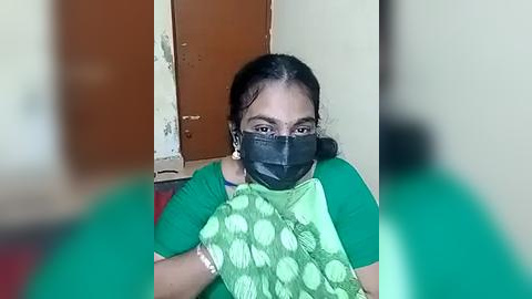 Media: Video of a woman with medium brown skin, wearing a green saree with white polka dots, black mask, and green blouse, sitting in a simple indoor room with a brown door and a white wall.