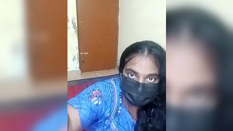 Media: A video of a South Asian woman with dark skin, long black hair, and brown eyes, wearing a blue floral blouse, black mask, and sitting on a red floor. The background shows a door and a wall.