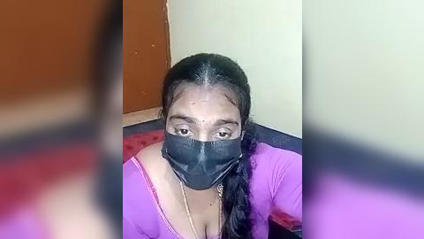 Media: Video of a woman with medium skin tone, black hair, wearing a black mask, pink shirt, sitting on a red carpet, indoors, blurred background.