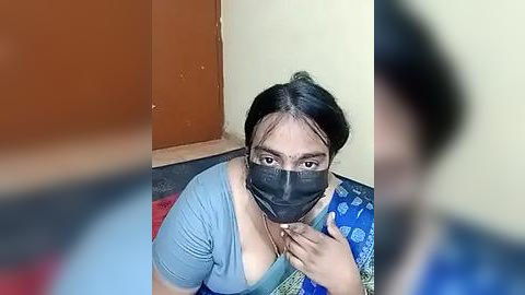 Media: A video of a South Asian woman with dark hair, wearing a blue top, black mask, and black face shield, kneeling on a red carpet, looking up.