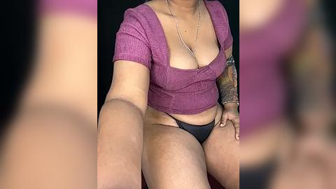 Media: A video of a plus-sized, light-skinned woman with a tattooed arm, wearing a low-cut purple top and black thong, taking a selfie. Background is out of focus.