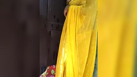 Media: Video of a person with light skin, wearing a yellow saree and holding a red, floral-patterned bag, standing in a dimly lit, concrete-walled corridor.