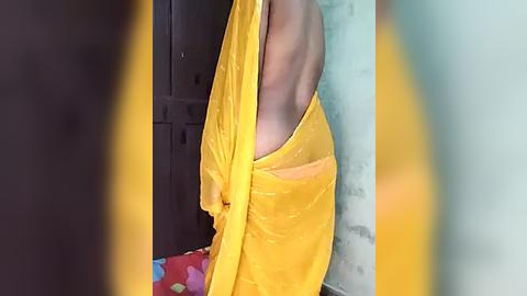 Media: Video of a person wearing a yellow saree, draped over the right shoulder, revealing the back. Background features a dark locker, colorful towel, and a white wall. The image is slightly blurry, with yellow and green borders.