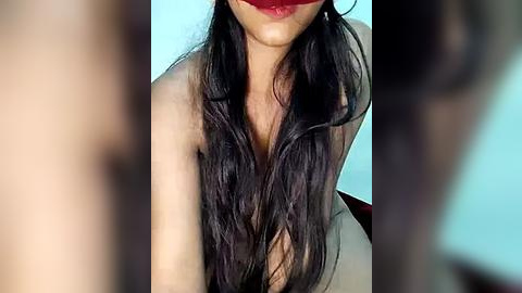 Media: A close-up video of a person with long, wavy dark hair, partially covering their face, wearing bold red lipstick. The background is blurred, featuring light blue and black hues.