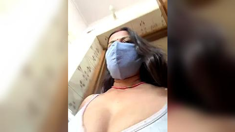 Media: Video of a woman with long dark hair, wearing a blue mask, white tank top, and red necklace, standing in a tiled room.