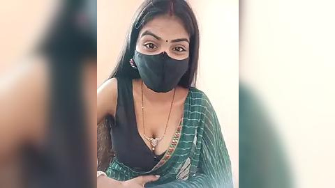 Media: Video of an Indian woman in a green saree with a black blouse, wearing a face mask and bindi, seated in a blurred indoor setting.