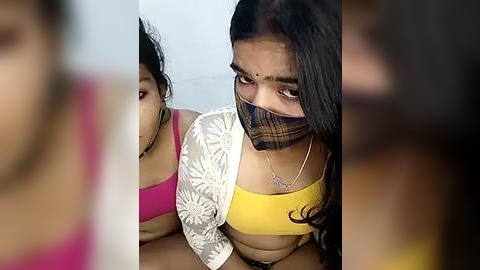 Media: Video of two women with medium skin tones, wearing traditional Indian attire, a yellow blouse and a pink bra, with a blurred background.