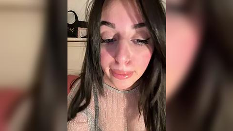 Media: A video of a young woman with long, dark hair, wearing a sheer, glittery top, sitting indoors. Her expression is neutral. The background shows a blurry, beige-colored wall and a kitchen countertop with objects.