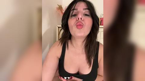 Media: A video of a young woman with light skin, dark hair, and large breasts, puckering her lips and wearing a black tank top, in a home setting with blurred figures in the background.