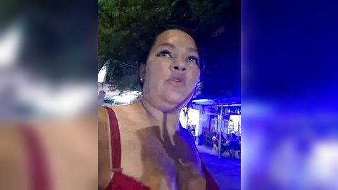 Media: Video of a woman with dark hair, wearing a red top, looking contemplative, standing outdoors at night with blurred lights in the background.