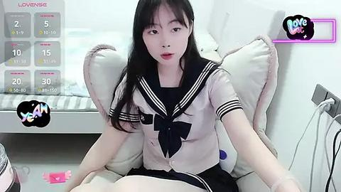 Media: A young East Asian woman with long black hair and fair skin, wearing a sailor school uniform, sits on a white chair in a well-lit room.