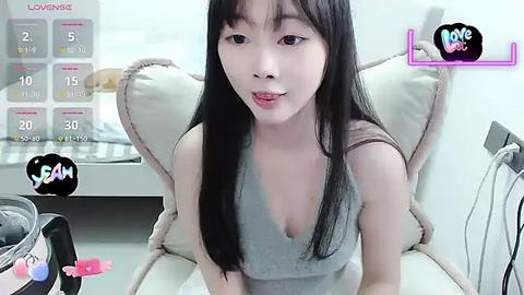 Media: A video of an Asian woman with long black hair, fair skin, and a slender build, wearing a grey tank top, sitting on a white armchair in a modern room.