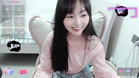 Media: A video of a smiling Asian woman with long black hair, wearing a light pink off-shoulder top and denim overalls, sitting on a beige chair in a modern room with white walls and a digital clock.