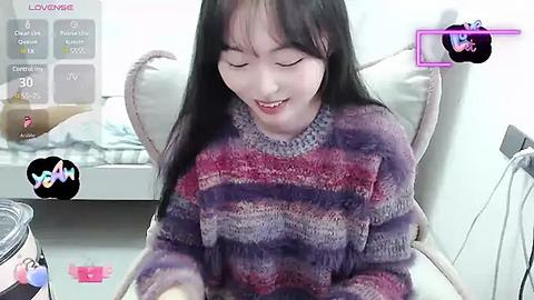 Media: Video of a smiling Asian woman with long black hair, wearing a multicolored striped sweater, sitting in a modern chair in a bright room.