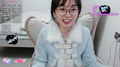 Media: Video of an East Asian woman with fair skin, glasses, and black hair, smiling, wearing a light blue furry jacket, sitting on a beige chair in a modern, minimalist bedroom with a bed in the background.