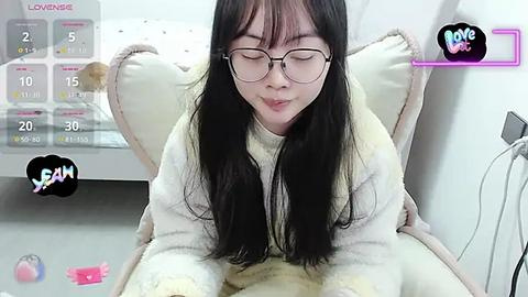 Media: A video of an East Asian woman with long black hair, wearing glasses, and a white fluffy robe, sitting on a beige cushion, in a cozy indoor setting.