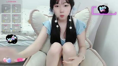 Media: A video of a young Asian woman with long black hair, sitting on a white chair, wearing a light blue top, and holding a pink vibrator. The background features a bed and a calendar with \"Love\" and \"Boy\" icons.