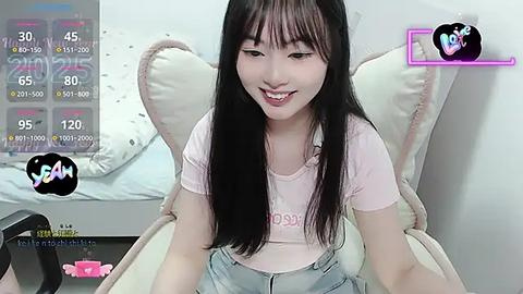 Media: A video of a young Asian woman with long black hair, wearing a pink T-shirt and denim overalls, sitting on a plush armchair in a bedroom.