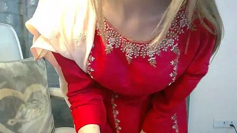 Media: Video of a woman in a red, embroidered Punjabi suit with gold floral patterns. She wears a beige dupatta draped over her shoulders. The background features a patterned cushion and a white wall.