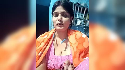 Media: Video of a South Asian woman with medium skin tone, dark hair, and a pink top, wearing a bright orange polka-dot shawl, in a sunlit room with a metal gate and a blurred orange object.