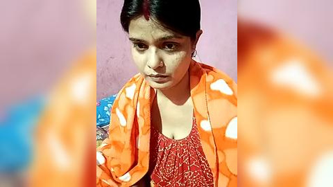 Media: Video of a young South Asian woman with dark hair, wearing a red blouse, orange polka-dot shawl, and blue sari. She has a serious expression, standing against a pink wall.