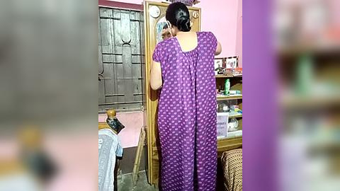 Media: A video of a woman in a purple polka-dotted dress, holding a phone, facing a mirror in a pink-walled room with a wooden door, shelves, and a woven basket.