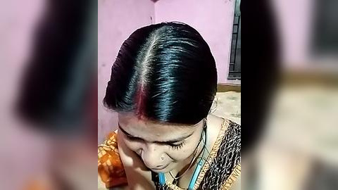 Media: Video of an Indian woman with black hair and a red bindi, wearing a traditional saree, sitting on a bed in a dimly lit room with pink walls.