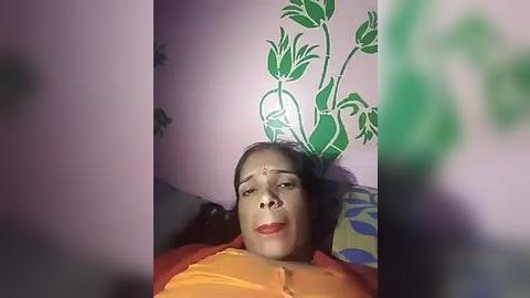 Media: Video of a middle-aged woman with medium skin tone, wearing a bright orange sari, lying on a bed with a floral-patterned wall behind her. She has a serene expression with red lipstick and a bindi on her forehead.