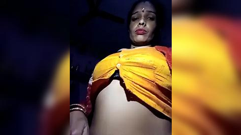 Media: Video of a confident woman with medium skin tone and black hair, wearing a bright yellow saree with a red border, and a red blouse, standing in a dimly lit room.