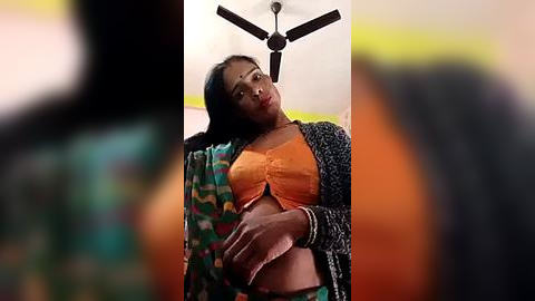 Media: Video of a South Asian woman with medium skin tone, long black hair, wearing a colorful saree, orange bra, and black cardigan, standing indoors with a ceiling fan above.