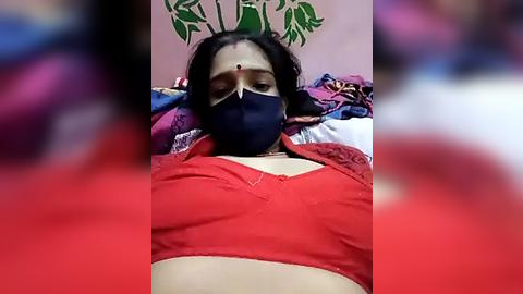 Media: Video of a woman lying on a bed with a red shirt and black mask, blurred background.