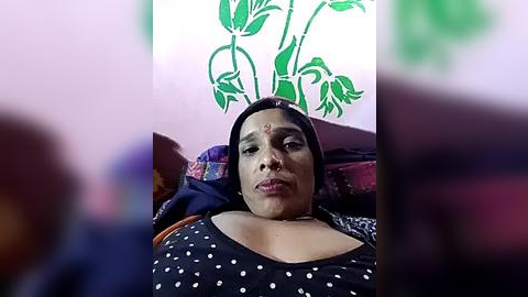 Media: Video of a woman with medium skin tone, large breasts, and a black polka-dotted dress, lying on a bed with a green leaf design wall behind her.