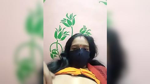 Media: Video of a woman with long black hair, wearing a black mask, orange scarf, and red blouse, sitting against a pink wall adorned with green lotus decals.